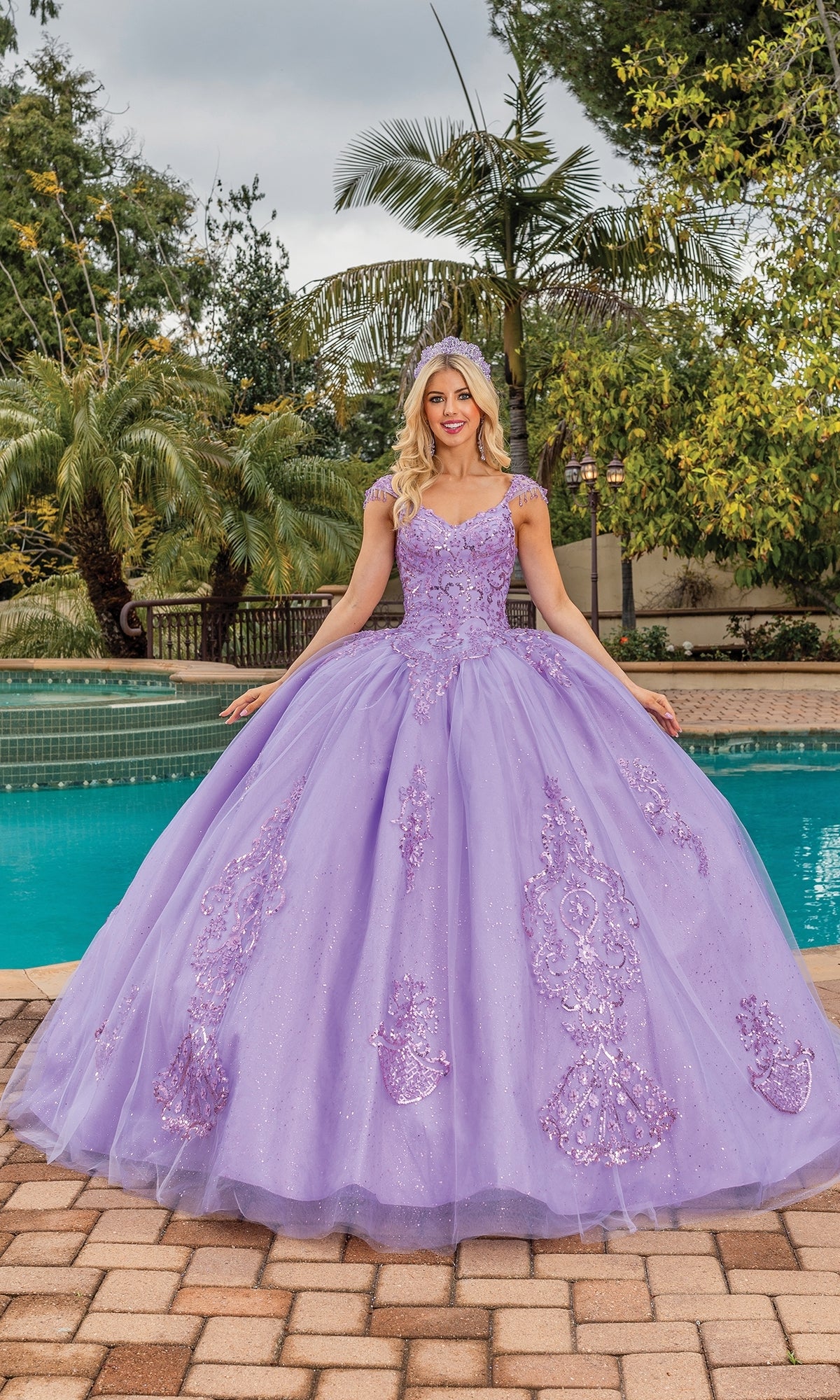 A woman in the Quinceanera Dress 1758 By Dancing Queen, featuring glitter lace embroidery and sequin details, stands on a brick patio in front of a pool and lush greenery.